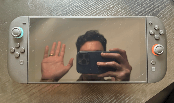 A picture of the Switch 2 with its screen turned off, bearing a reflection of me taking a picture of it, waving my hand hello.